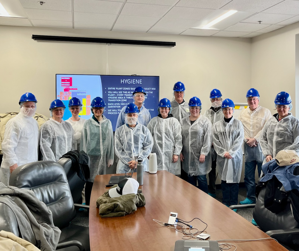 A large group of people in blue hard hats and see-through lab coats at the end of a large conference table.