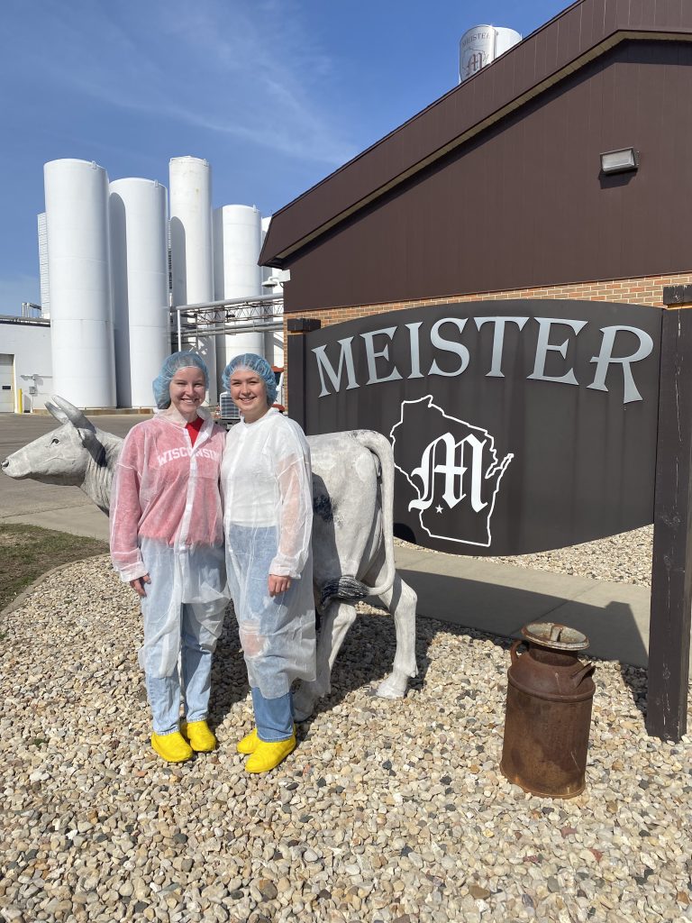 Two people in lab coats and hairnets stand in front of cheese plant sign, with processing facilities in the background.